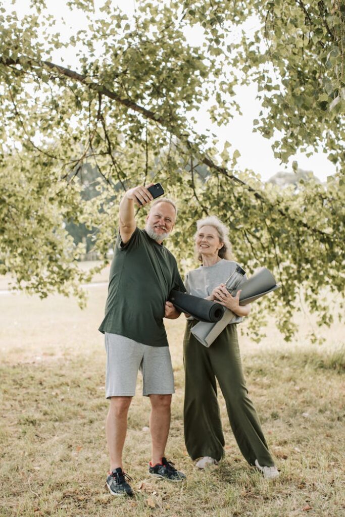 Elderly couple taking a selfie in a park, holding yoga mats, enjoying outdoor fitness.