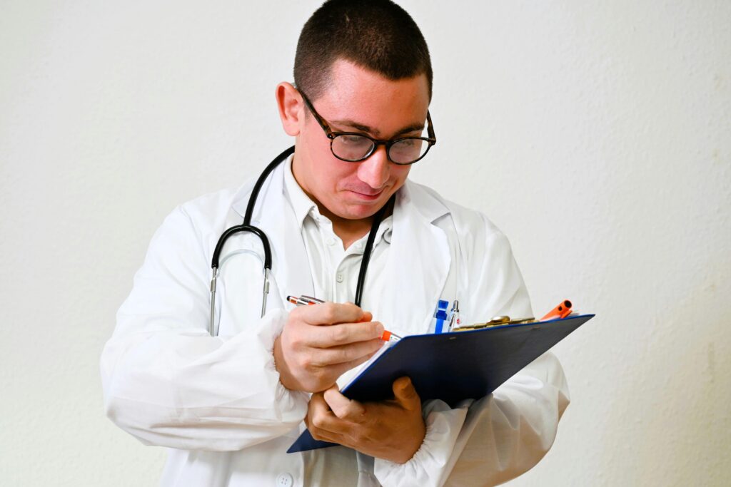 A male doctor in a white coat writes on a clipboard in a hospital setting, showcasing healthcare professionalism.