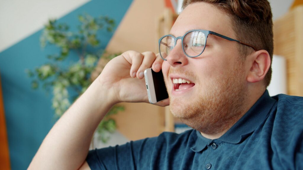 Happy adult man with glasses having a phone conversation inside a bright room.