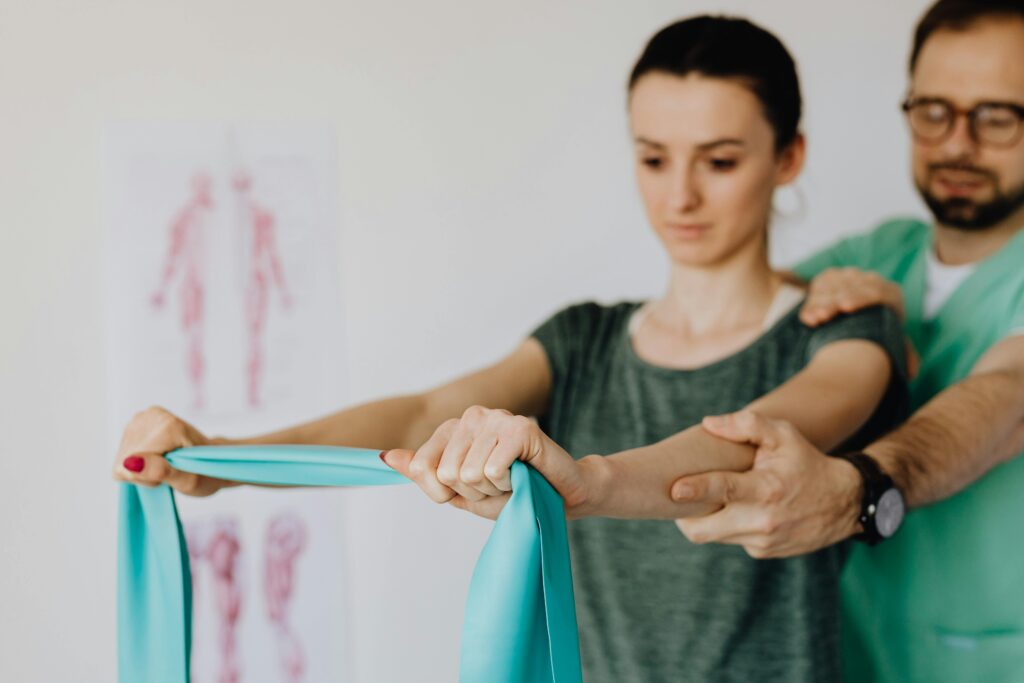 Crop osteopath in uniform and eyeglasses supporting shoulder and reached arm of young woman with elastic tape in doctor office in hospital on blurred background