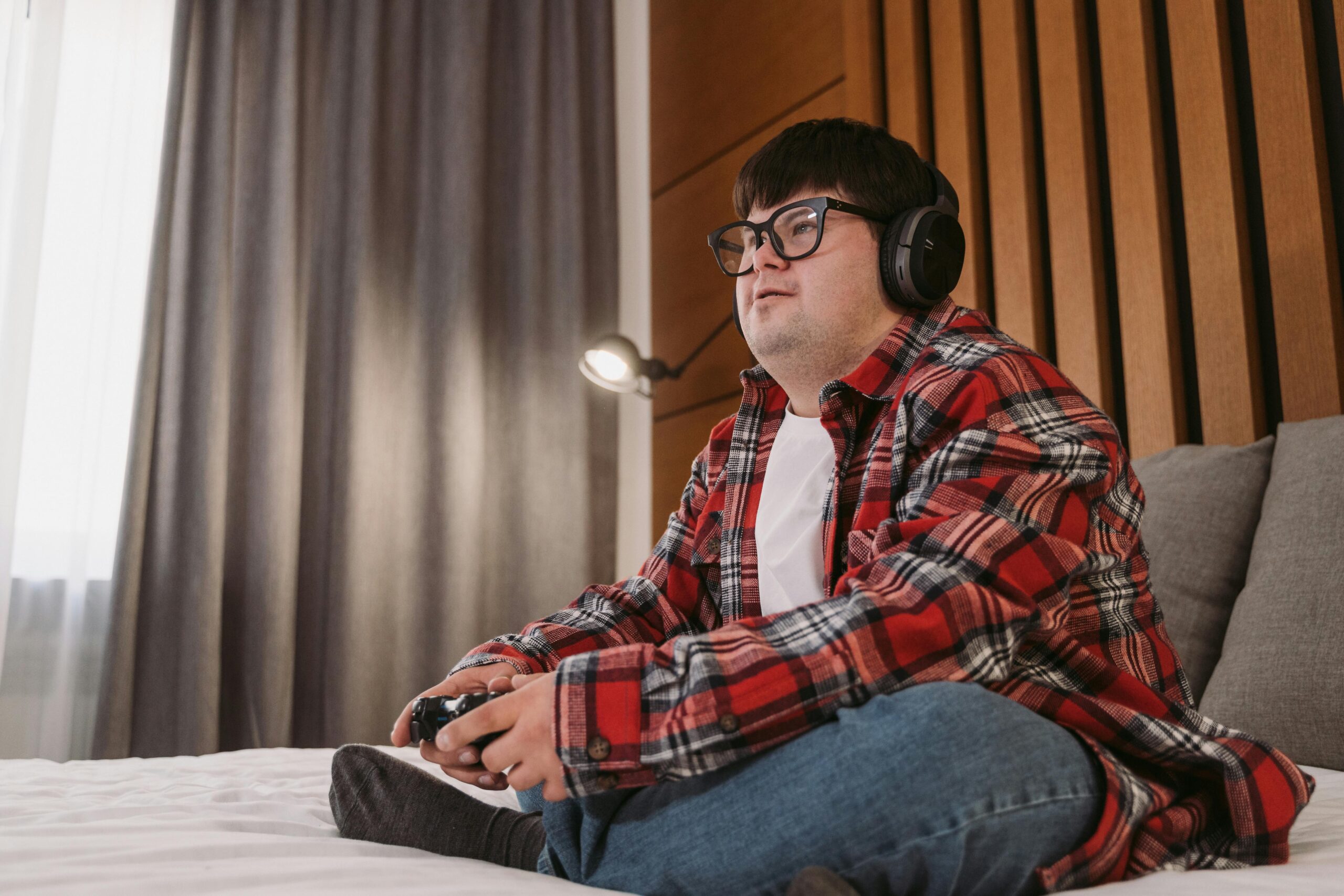 Young man with headphones playing video games on a bed, enjoying leisure time indoors.