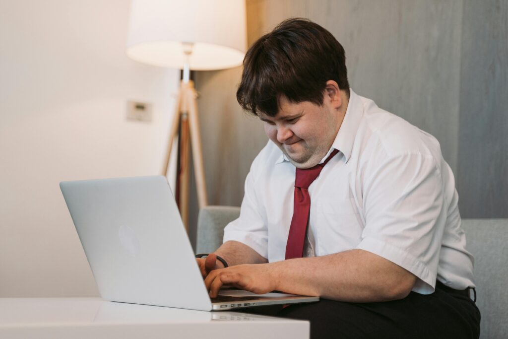 A young man with Down syndrome smiling while working on a laptop indoors.