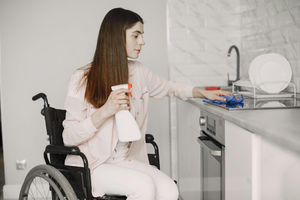 A woman in a wheelchair cleaning a modern kitchen countertop using a spray bottle and cloth.