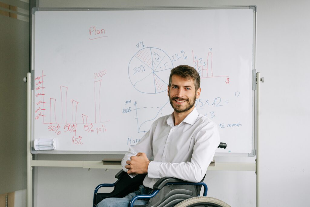 A smiling man in a wheelchair giving a business presentation with charts on a whiteboard.