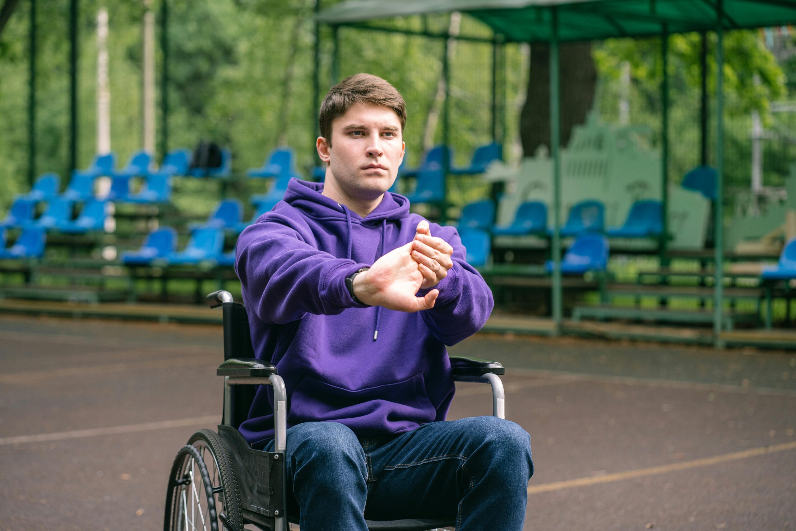 Adult man in a purple hoodie stretching his arms while sitting in a wheelchair outdoors.