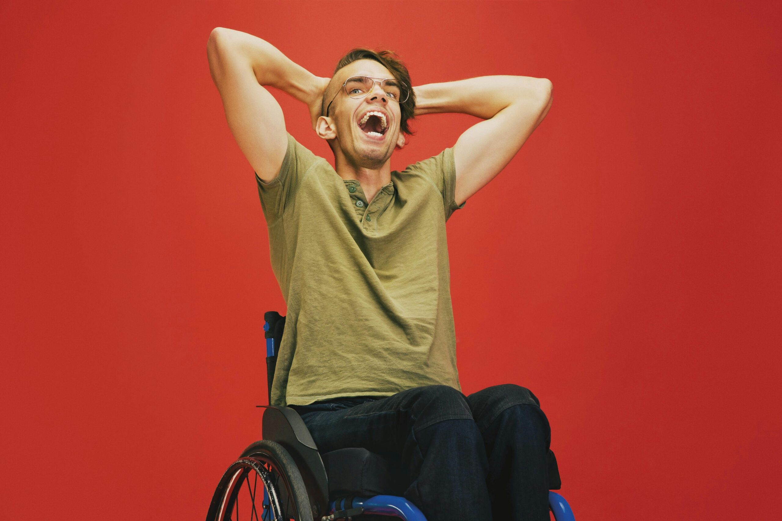 Smiling man in a wheelchair with hands on head, against a vibrant red backdrop.